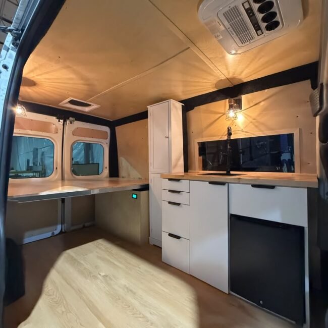 Camper van interior with custom white cabinetry, sink, fridge and rooftop air conditioning.