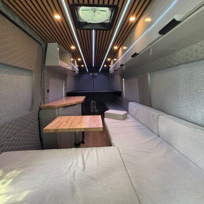 L-shaped camper lounge with gray cushions, butcher-block table, custom cabinets and illuminated slatted wood ceiling.