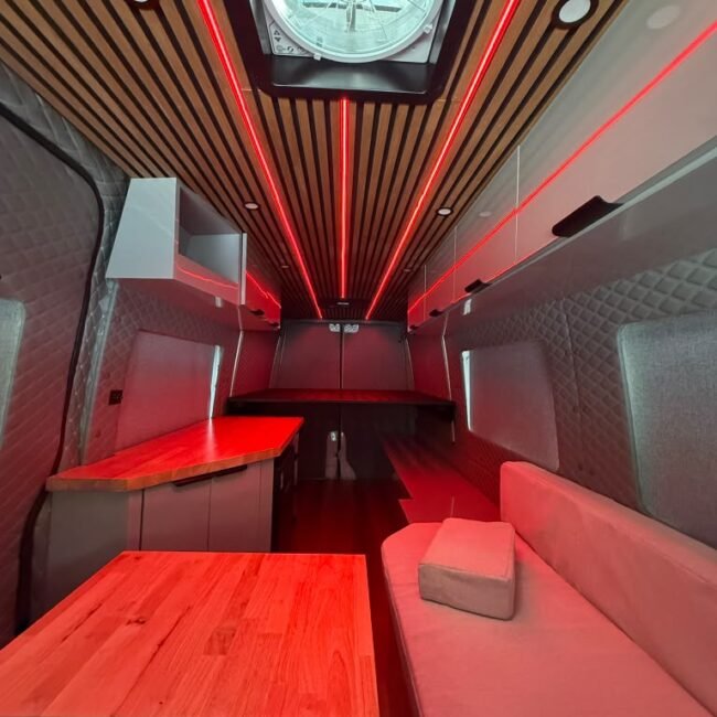 Custom camper interior with L-shaped lounge, wood surfaces, slatted ceiling and red LED accent lighting.