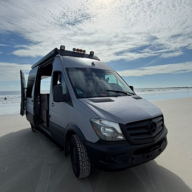 Front exterior view of the Mercedes Sprinter camper on the beach with roof-mounted auxiliary lights.